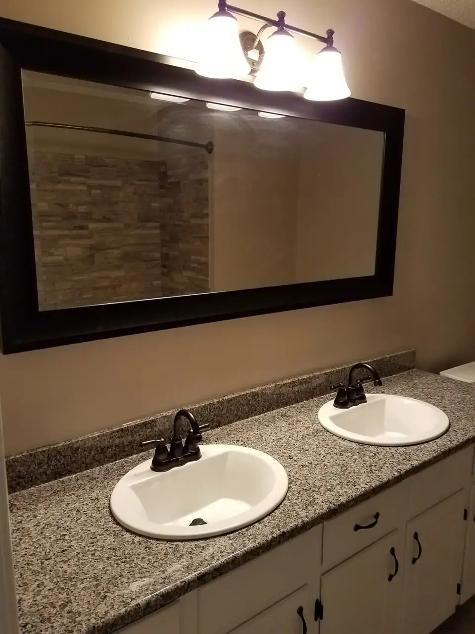 Bathroom double vanity with a granite top, two white sinks, black faucets and a framed mirror, part of L H Carpentry's remodeling in Nacogdoches, TX